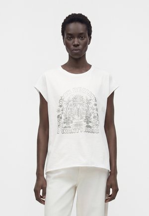 Femme aux cheveux courts et bouclés porte un haut blanc sans manches et un pantalon, le haut présente un motif illustré et le texte « JE VEUX DE LA MAGIE. »