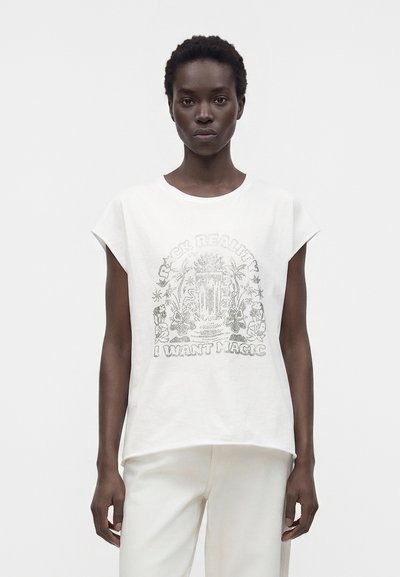 Femme aux cheveux courts et bouclés porte un haut blanc sans manches et un pantalon, le haut présente un motif illustré et le texte « JE VEUX DE LA MAGIE. »