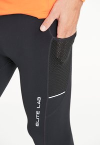 Main masculine reposant à l'intérieur d'une poche en maille noire sur des leggings de sport noirs avec le texte « ELITE LAB » et un t-shirt orange visible.