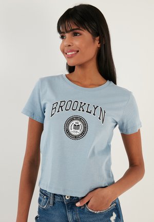 Heled sinise värviga lühikeste varrukatega t-särk, mis on valmistatud puuvillast. Esiküljel on mustade tähtedega "BROOKLYN" ning ringikujuline logo, mis sisaldab teksti ja krooni disaini.