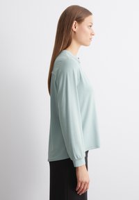 Top a maniche lunghe di colore verde menta in tessuto morbido, caratterizzato da una vestibilità ampia, scollo rotondo e polsini arricciati, abbinato a un pantalone nero.