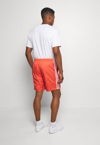 Pantaloni sportivi da uomo in vivace corallo con strisce laterali bianche, abbinati a una t-shirt bianca semplice e sneakers bianche.