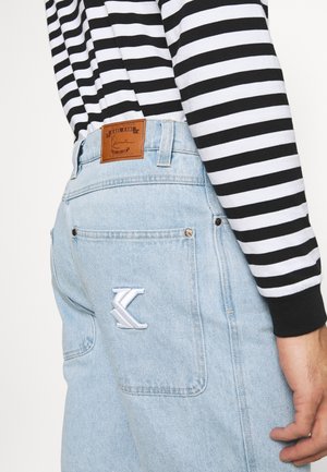 Jean en denim bleu clair avec une poche arrière ornée d'un logo "K" blanc brodé, porté avec un t-shirt à manches longues rayé noir et blanc.