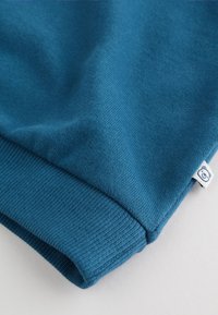 Sweat-shirt bleu sarcelle avec un bord-côte au poignet et une petite étiquette blanche présentant un logo d'ours. Texture douce avec une finition lisse et impeccable.