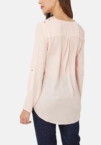 Mujer vestida con una blusa rosa clara de mangas largas, con detalles de botones en los hombros y brazos, combinada con pantalones vaqueros azul oscuro, vista desde atrás.