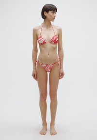 Conjunto de biquíni com padrões florais vermelhos sobre fundo branco. Top triangular com alças ajustáveis e calções com laços laterais. Textura de tecido suave.