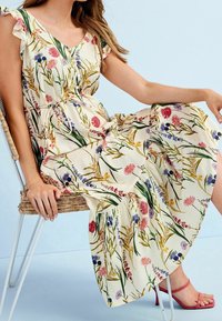 Bloemige maxi-jurk met korte, gerimpelde mouwen, met een crèmekleurige achtergrond, kleurrijke bloemen, een strikceintuur en een vloeiende rok. Roze sandaaltjes met hak.