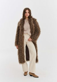 Žena v dlouhém hnědém chlupatém kabátu, béžovém svetru, krémových širokých kalhotách a hnědých kotníkových botách stojí před jednobarevným světlým pozadím.