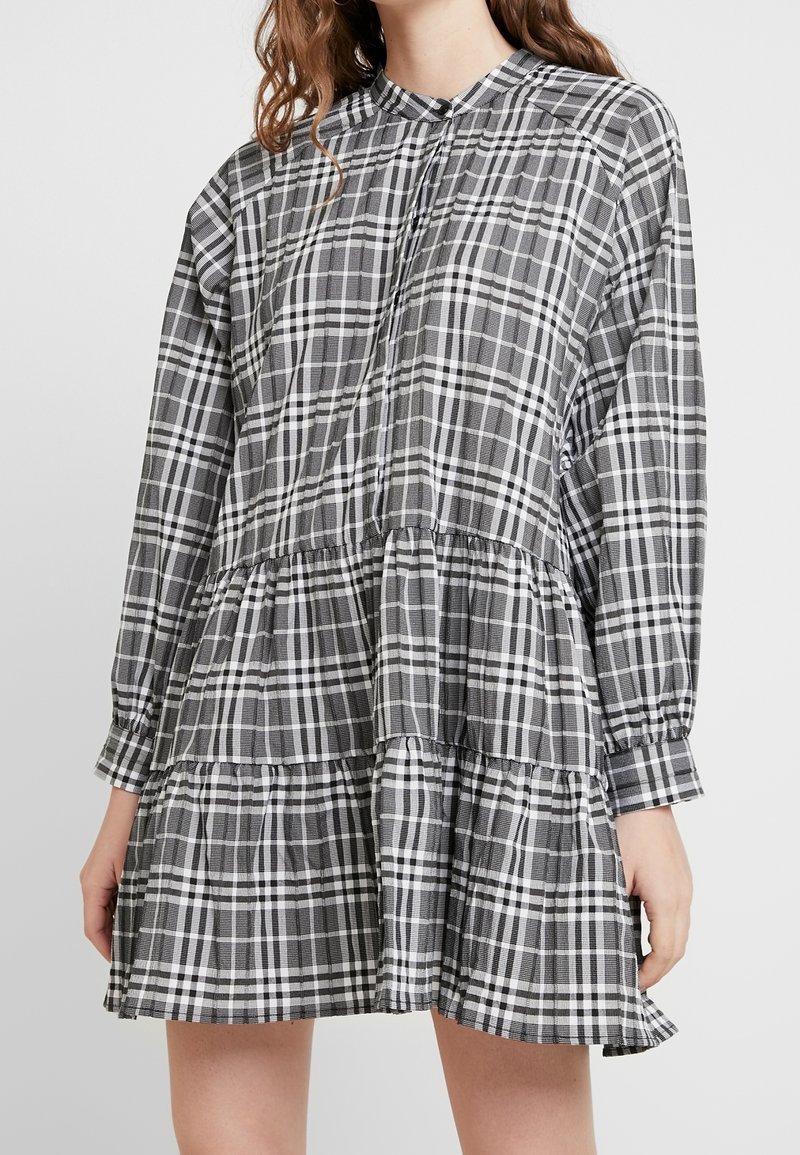 Femme portant une robe à manches longues à carreaux noir et blanc, à volants, avec un col montant et des poignets boutonnés.