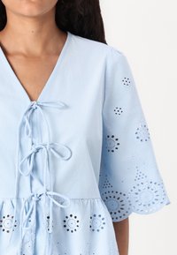 Top di cotone azzurro chiaro con scollo a V, chiusura con lacci sul davanti e maniche corte, caratterizzato da ricami a occhiello decorativi e dettagli con bordo scallop.
