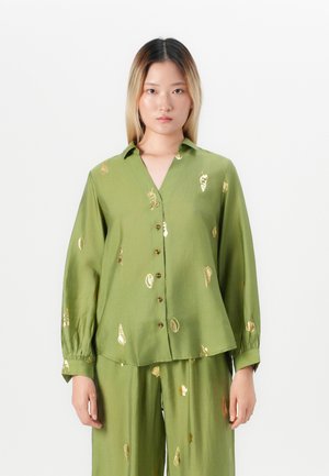 Donna che indossa una camicia verde oliva con bottoni e pantaloni abbinati con motivi dorati a conchiglie, in piedi davanti a uno sfondo bianco semplice.