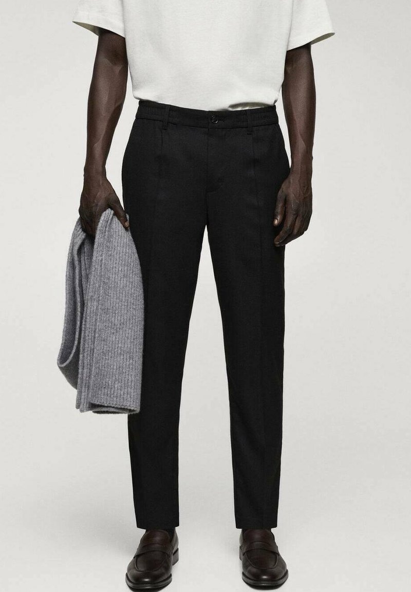 Homme portant un pantalon noir, des mocassins marron foncé, une chemise blanche à manches courtes, tenant un pull gris en maille plié dans la main gauche.