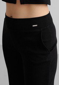 Svart ribbade byxor med bred elastisk midjeband och sidoficka, med en liten silverfärgad logoprydnad nära midjebandet.