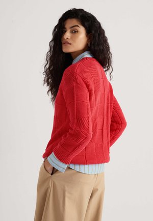 Donna con capelli lunghi e ricci che indossa un maglione rosso a maglia lavorata sopra una camicia a righe blu e pantaloni beige, con le mani nelle tasche, che guarda oltre la spalla.