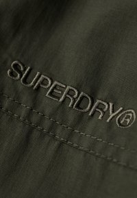 Grøn stof med en tekstureret overflade, der har syede detaljer. "SUPERDRY" logoet er broderet i en lysere grøn nuance.