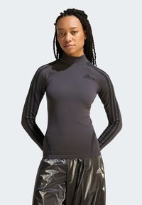 Žena s pletenými vlasmi, ktorá má na sebe priliehavý čierny dlhý rukáv značky Adidas a lesklé čierne nohavice Adidas, stojaca s rukami za chrbtom.