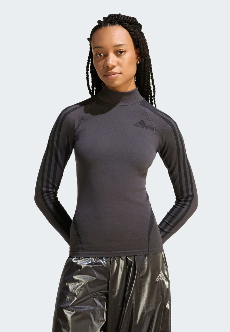 Žena s pletenými vlasmi, ktorá má na sebe priliehavý čierny dlhý rukáv značky Adidas a lesklé čierne nohavice Adidas, stojaca s rukami za chrbtom.