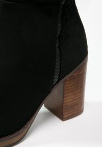 Bottine noire à cheville présentant une texture en daim lisse, un détail tressé sur le côté, et un talon bloc en bois avec des rainures pour une meilleure adhérence.