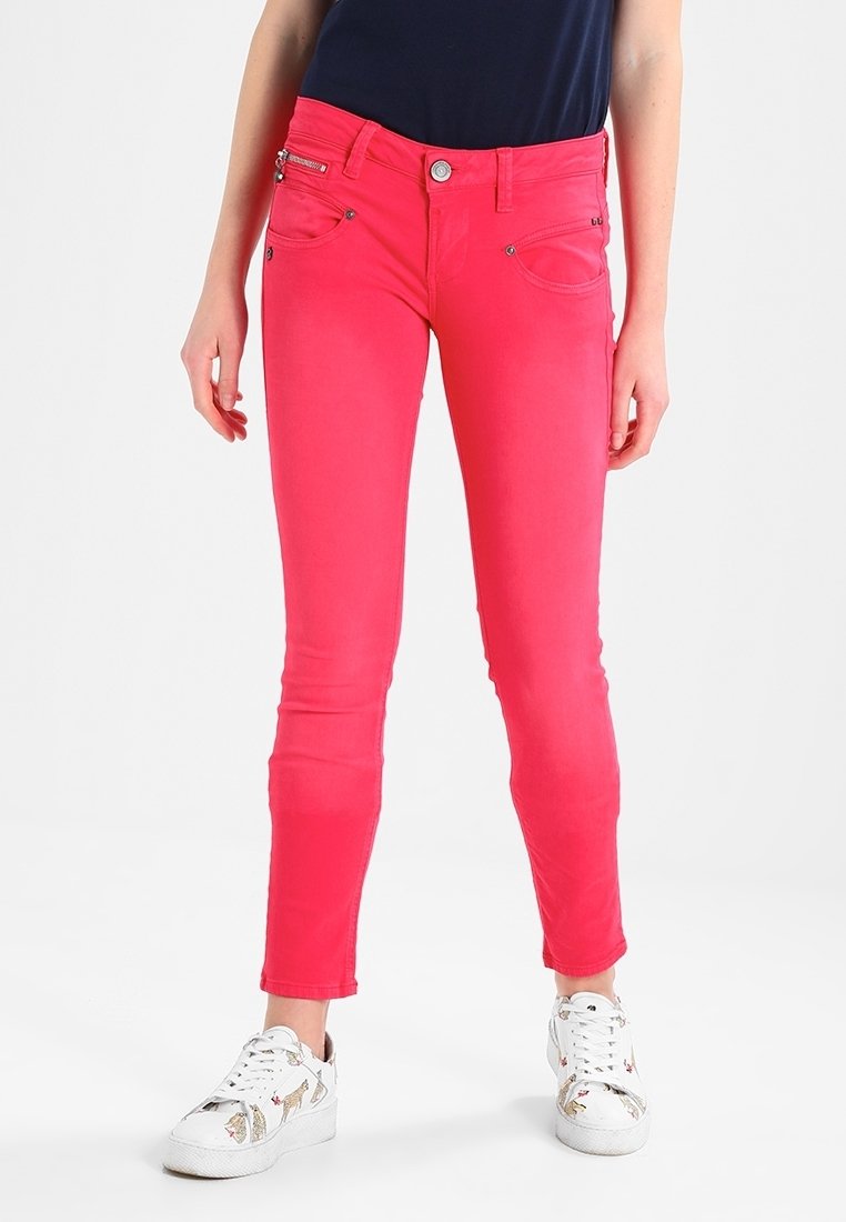 Mujer con pantalones ajustados rosa brillante, camiseta azul marino y zapatillas blancas con estampado animal, de pie sobre un fondo blanco.