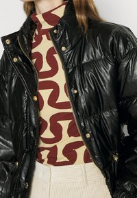 Czarna pikowana kurtka z imitacji skóry z złotymi akcentami, zamkiem błyskawicznym i wysokim kołnierzem, noszona na wzorzystym golfie.