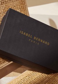 Boîte rectangulaire noire avec une surface texturée, affichant du texte doré en relief "ISABEL BERNARD" et "PARIS" sur le dessus.