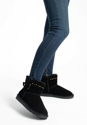 Bottines en suède noir avec une doublure en fausse fourrure douce, dotées de clous dorés et d'une sangle, associées à un jean skinny bleu foncé.