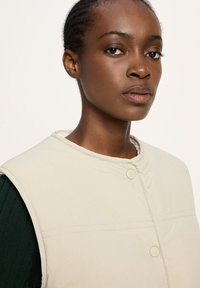 Beige Puffer-Weste mit rundem Ausschnitt und Knopfverschluss, präsentiert an einem Modell mit strukturiertem Haar. Glatte Stoffoberfläche und minimale Nähdetails.