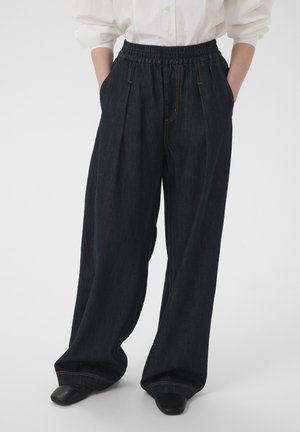 Persoon die een donker denim broek met wijde pijpen en een elastische tailleband draagt, handen in de zakken, gecombineerd met zwarte schoenen en een wit ingestoken overhemd.