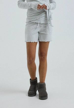 Persona que lleva pantalones cortos gris claro y una camiseta a juego con agujeros para los dedos, de pie con botines gris oscuro sobre un fondo claro y sencillo.