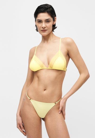 Γυναίκα με κοντά σκούρα μαλλιά που φοράει ένα κίτρινο τοπ μπικίνι με δαντέλα και ταίριαστο κάτω μέρος, μπροστά σε απλό ανοιχτό φόντο.