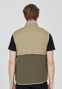 Beige och olivgrön ärmlös väst med en slät överdel, texturerad fleece-nedre del och ljusfärgad kant, som har en hög krage.