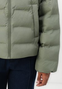 Olijfgroen gewatteerde jas met een rits aan de voorkant, een gestikte uitstraling en een zijzak, gecombineerd met donkerdenim broek. Glad, matte textuur.