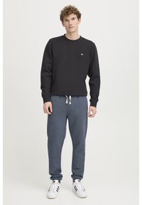 Sort sweatshirt med rund hals og lille hvidt logo, kombineret med mørkegrå joggers med snørre, afsluttet med hvide sneakers.