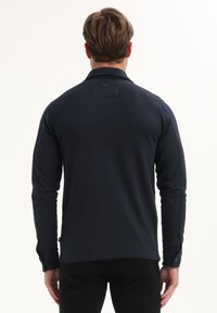 Hombre con cabello castaño claro que lleva una camisa de manga larga azul marino y pantalones negros, visto de espaldas contra un fondo blanco liso.