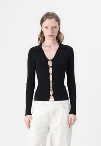 Zwarte blouse met lange mouwen en een V-hals, voorzien van een rij ovale knopen met metalen accenten aan de voorkant, gecombineerd met een witte broek.