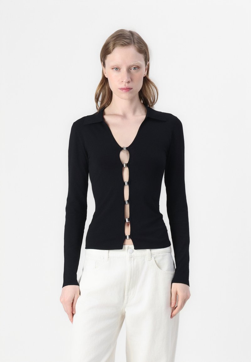 Zwarte blouse met lange mouwen en een V-hals, voorzien van een rij ovale knopen met metalen accenten aan de voorkant, gecombineerd met een witte broek.
