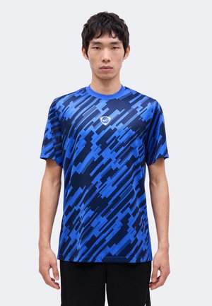 Jeune homme debout face à l'avant, portant un t-shirt à manches courtes bleu et noir à motifs avec un petit logo sur la poitrine.