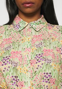 Camisa de botones floral multicolor con un fondo beige claro, cuello corto y patrones vibrantes de flores en rosa, púrpura y amarillo.
