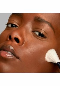 Nahaufnahme eines glatten, bronzefarbenen Gesichts mit glänzenden Lippen. Ein weißer, weicher Makeup-Pinsel trägt Produkt auf die Wange auf. Die natürliche Hautstruktur ist sichtbar.