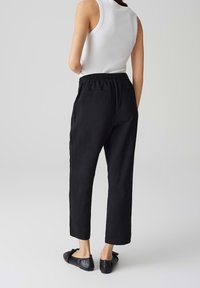 Pantalons noirs à taille élastique avec une coupe loose et droite, fabriqués dans un tissu texturé. Portés avec un haut sans manches blanc et des chaussures plates noires.