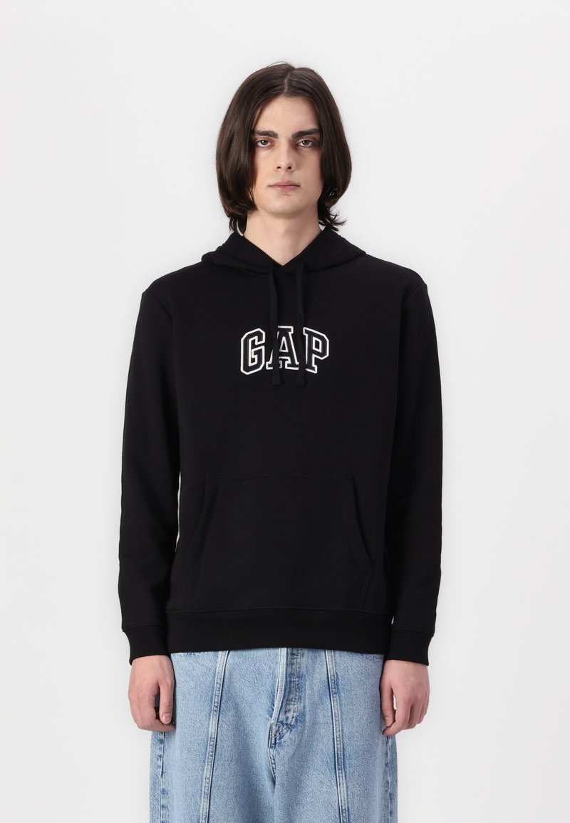 Hoodie en coton noir avec une poche kangourou à l'avant et le logo "GAP" en blanc. Comprend une capuche avec cordon de serrage et des poignets côtelés. Porté avec un jean large bleu clair.