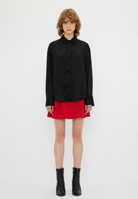 Camicia nera a maniche lunghe con colletto, abbinata a una gonna a ruota rossa brillante. L'outfit è completato da stivaletti neri alla caviglia.