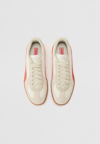 Tenisi bej cu un amestec de textil neted și suede, cu accente roșii, sireturi și o talpă din cauciuc de tip gumă. Logo Puma pe limbă.