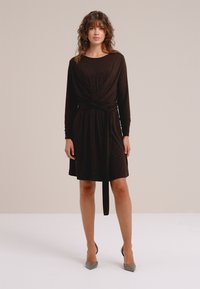 Vrouw die een zwarte jurk draagt met lange mouwen en knielengte, met een ceintuur in de taille en grijze pumps met een spits toelopende neus, staand tegen een effen achtergrond.