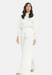 Blusa bianca con colletto alto e chiusura a bottoni abbinata a pantaloni a vita alta e gamba larga dello stesso colore. Tessuto liscio e lucido con sottili fantasie a pois.