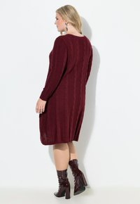 Donna con capelli biondi che indossa un vestito midi in maglia a trecce color bordeaux e stivaletti alla caviglia in pelle verniciata con tacco quadrato, rivolta a sinistra.