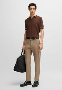 Brązowa koszulka polo z krótkim rękawem i białymi wykończeniami, beżowe dopasowane spodnie, czarny pasek i buty, trzyma czarną skórzaną torbę. Prosty, nowoczesny styl.