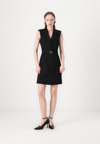 Black sleeveless dress with a belted waist, tailored fit, and lapel collar. Model wearing pointed-toe black heels. Smooth texture. Plain background.