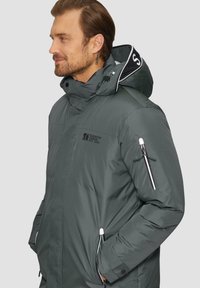 Dunkelgrüne isolierte Jacke mit hohem Kragen, verstellbarem Kapuze, Seitentaschen und einer mit Reißverschluss versehenen Ärmeltasche. Mit Logo und kontrastierendem Besatz.