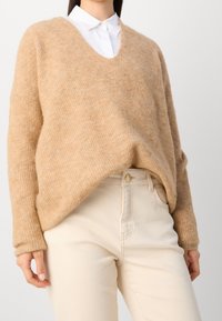Bežový pletený svetr s výstřihem do V, volným střihem a žebrovanou texturou, nošený přes bílou košilku s límečkem a spárovaný se světle bežovými džíny.